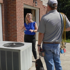 HVAC technician speaking with homeowner beside outdoor air conditioning unit outside a brick house.