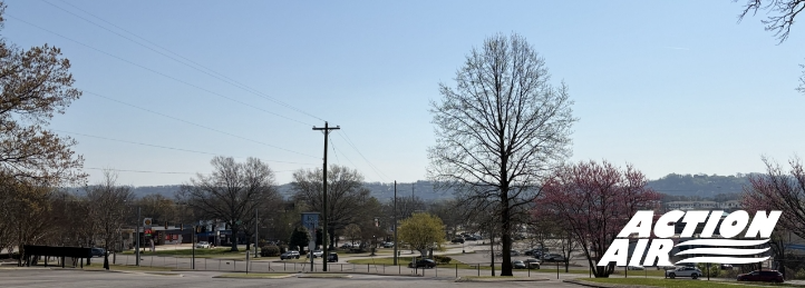 Wide view of a small town street with trees, parked cars, and rolling hills in the background.