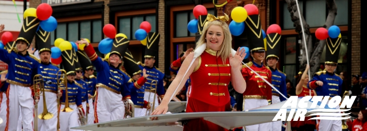 Parade performer in red costume waving from float with marching band and colorful balloons in the background.